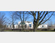 Das Bild zeigt das Gebäude des Bundeskanzleramts in Berlin, aufgenommen aus einem Park mit kahlen Bäumen im Vordergrund. Die moderne Architektur des Gebäudes hebt sich durch große Glasflächen und helle Fassaden hervor, während der blaue Himmel die Szene dominiert.