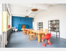 Großer, heller Raum mit modernen, schlichten Möbeln; ein langer Holztisch mit Stühlen und einem roten Kunststoffstuhl im Vordergrund. Eine blaue Wand im Hintergrund enthält ein Regal, daneben stehen Glasvitrinen und ein Sessel an der weißen Wand.