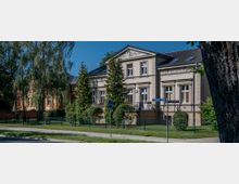 Helles, zweistöckiges Gebäude im klassischen Stil mit dekorativen Fassadenelementen und Satteldach, umgeben von Bäumen, Sträuchern und einem Zaun. Im Vordergrund befindet sich ein Straßenschild mit der Beschriftung „Fehrbelliner Straße“.