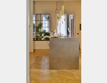 Moderner Innenraum mit einem zentralen, metallischen Tresen, darüber mehrere Pendelleuchten mit goldenen und gläsernen Elementen. Im Hintergrund ist ein großes Fenster mit schwarzen Metallrahmen zu sehen, durch das Pflanzen sichtbar sind, sowie ein heller Raum mit Holzparkettboden.