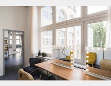 Ein heller Raum mit großen Fenstern, die Ausblick auf Außenfassaden und Bäume bieten. Im Vordergrund steht ein Holztisch mit einem beleuchteten Schminkspiegel; im Hintergrund ist ein weiterer Raum mit Regalen und einem offenen Grundriss sichtbar.