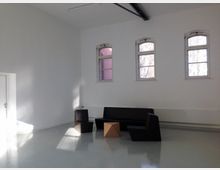 Ein minimalistischer, heller Raum mit weißen Wänden und glänzendem, grauem Boden. Drei schmale Fenster mit Rundbögen befinden sich an der Wand, und im Raum stehen eine dunkle Sitzbank sowie geometrische Holzhocker.