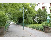 Gepflasterter Gehweg in einer grünen Parkanlage, umgeben von Bäumen und Sträuchern, mit grünen Straßenlaternen und einem angrenzenden Zaun. Rechts sind ein Hochhaus und ein Verkehrsschild zu sehen, das einen Weg für Fußgänger und Radfahrer kennzeichnet.