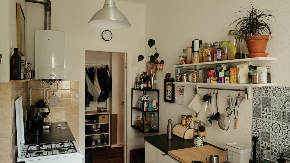 Kleine Wohnküche mit Gasherd, Arbeitsplatte und Spüle, über der offene Regale mit Vorratsgläsern, Gewürzen und hängenden Küchenutensilien montiert sind. Weiße Wände, eine große Hängelampe, gemusterte Fliesen an der Spüle sowie ein Durchgang zu einem Flur mit Garderobe prägen den Raum.