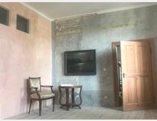 Ein schlicht eingerichteter Innenraum mit einer teilweise verputzten, teils alt wirkenden Wand, einem holzgerahmten Gemälde, einem offenen Holztürrahmen und einem kleinen Set aus antikem Stuhl und Beistelltisch. Der Boden besteht aus hellen Holzdielen.