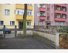 Mehrfamilienhaus mit gelber und rosa Fassade, Balkonen und großen Fenstern, umgeben von einem Metallzaun und einem kleinen, begrenzten Hofbereich mit Baum und kahlen Sträuchern; am Rand geparkte Autos.