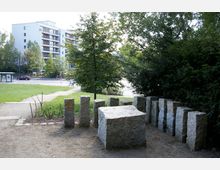 Eine kleine Grünfläche mit einer Anordnung von aufrechten Granitsteinen und einem flachen Steinblock im Vordergrund. Im Hintergrund sind ein Bürgersteig, eine Straße, ein Haltestellenhäuschen, Bäume und ein modernes Wohngebäude zu sehen.