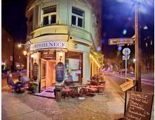Straßenszene bei Nacht mit dem Eckrestaurant "Sophieneck" in einem hell beleuchteten, historischen Gebäude. Vor dem Restaurant stehen Tische und Stühle auf dem Gehweg, während ein Straßenschild mit der Aufschrift „Sophienstraße“ und eine beleuchtete Kreidetafel mit Speisenangebot sichtbar sind.