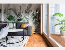 Wohnzimmer mit Holzboden und großem Fenster, das auf eine Terrasse führt. Das Interieur umfasst eine graue Couch, einen weißen Couchtisch, eine Stehlampe und eine Wand mit einem Pflanzenmotiv sowie Dekoration mit grünen Zimmerpflanzen.