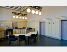 Ein historischer Sitzungsraum mit gelben Wandvertäfelungen, einer Decke mit Kassettendesign und einer dekorativen Hängelampe mit mehreren runden Leuchten. In der Mitte des Raumes steht ein trapezförmiger Tisch mit Stühlen; im Hintergrund befindet sich eine erhöhte Kanzel mit Holzmöbeln und geschlossenen Türen.