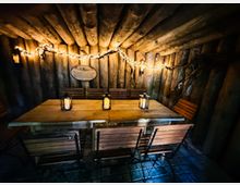 Rustikaler Holzkellerraum mit niedriger Decke aus Baumstämmen und Wänden aus grob behauenen Bohlen, beleuchtet von einer Lichterkette. In der Mitte steht ein großer Holztisch mit mehreren Stühlen und Laternen, der Boden ist mit dunklen Steinfliesen ausgelegt.