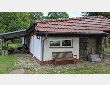 Kleines Gebäude mit rotem Ziegeldach und weißer Fassade, daneben eine Steinwand mit alter Holztür. Im Vordergrund steht eine Holzbank auf einem gepflasterten Weg, umgeben von grünem Gras und Bäumen im Hintergrund.