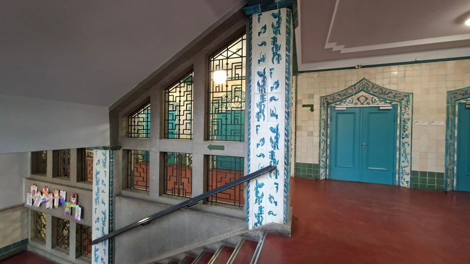 Innenansicht eines Treppenhauses mit breiten Fenstern in geometrischen Sprossenmustern und einem Treppengeländer entlang der Stufen. An den Wänden und Säulen sind helle Fliesen mit blau-weißen Ornamenten, gegenüber stehen türkisfarbene Doppeltüren mit dekorativ gerahmtem Türbogen.