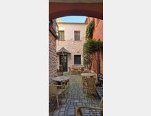 Kleiner Innenhof mit Kopfsteinpflaster und mehreren runden Café-Tischen mit Korbstühlen, umgeben von Naturstein- und rot verputzten Wänden. Im Hintergrund stehen eine grüne Eingangstür mit kleinem Vordach und Fenster; an der rechten Wand wachsen Kletterpflanzen und ein kleiner Baum.