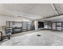 Große, leere Garage oder Lagerraum mit Betonboden und niedriger Decke, ausgestattet mit Metallregalen, Werkbank und Schränken entlang der Wände. An der rechten Seite befindet sich ein breites Tor, in der Decke verlaufen sichtbare Leitungen und Leuchten.