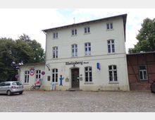 Historisches Bahnhofsgebäude in Rheinsberg (Mark) mit hellem Putz, mehreren Sprossenfenstern und einem Eingangsbereich mit einem Schriftzug über der Tür. Vor dem Gebäude befindet sich ein mit Kopfsteinpflaster ausgelegter Parkplatz, an dessen Rand Bäume stehen.