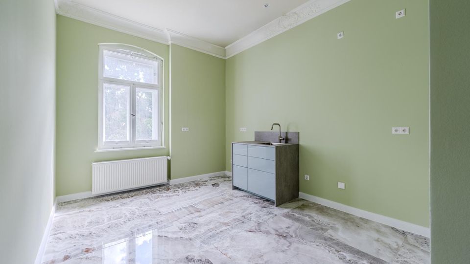 Leerer Raum mit hellgrünen Wänden, dekorativer Stuckleiste an der Decke und glänzendem, marmoriertem Fliesenboden. Links ein großes Fenster mit Heizkörper darunter, rechts ein kleiner Küchenunterschrank mit Spüle und mehreren Steckdosen an der Wand.