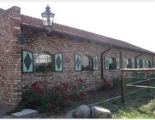 Langes, niedriges Gebäude aus roten Ziegelsteinen mit grünen Fensterläden, die ein Rautenmuster aufweisen. Davor befinden sich ein mit roten Blumen bepflanztes Beet und eine Laterne an der Wand, während im Vordergrund ein einfacher Holzzaun sichtbar ist.