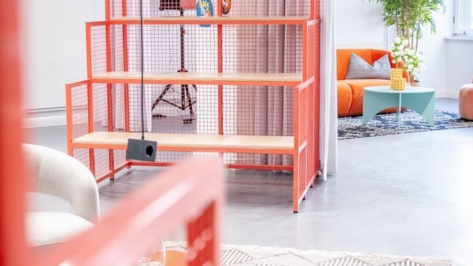 Heller, moderner Loft-Innenraum mit hohen Decken, weißen Wänden und Vorhängen sowie sichtbaren Installationen an der Decke. In der Mitte steht ein rotes Metallregal mit Holzablagen; daneben ein Sitzbereich mit orangefarbenem Sofa, blauem Couchtisch, Teppich, Hocker und einer großen Zimmerpflanze unter Pendelleuchten.