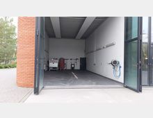 Offene Garage mit großer Glastür, weiß gefliesten Wänden und grauem Boden. Im Inneren befinden sich Reinigungsgeräte, Aufbewahrungsregale, ein Wasserschlauch an der Wand und eine abgesenkte Ablaufrinne auf dem Boden.