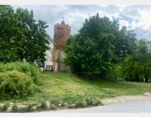 Historischer Rundturm aus rotem Backstein und Feldsteinfuß, umgeben von Bäumen und einer Grünfläche; daneben ein modernes Gebäude mit heller Fassade.