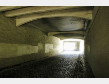 Ein gepflasterter Tunnel mit abgenutzten Wänden und freiliegenden Betonträgern an der Decke, der zu einem hell beleuchteten Ausgang führt.