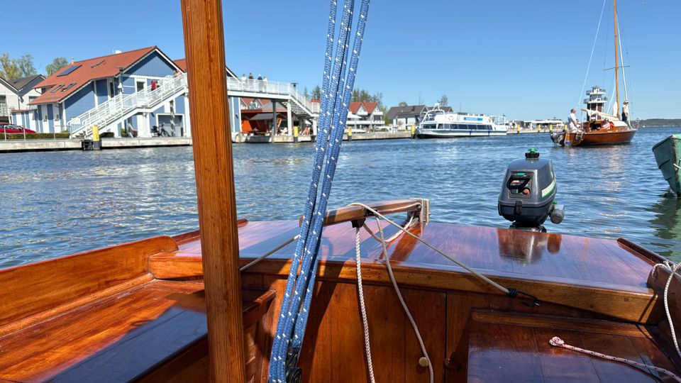 Blick vom hölzernen Deck eines kleinen Segelbootes über einen Hafenkanal, mit sichtbaren Leinen und Mast im Vordergrund. Am Ufer stehen Gebäude mit roten Dächern und Steganlagen, im Wasser liegen weitere Boote vor blauem Himmel.