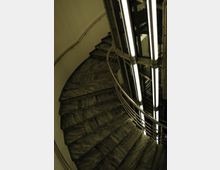 Spiralförmiges Treppenhaus mit dunkel gemaserten Marmorstufen und einem zentralen Aufzugsschacht mit vertikalen Leuchtstofflampen; das Geländer besteht aus Metall.