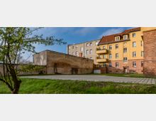 Hinterhof mit gepflastertem Weg und Rasenfläche, umgeben von unterschiedlichen Gebäuden, darunter eine gelbe, mehrstöckige Wohnhausfassade mit roten Ziegeldetails und ein verputztes Gebäude mit sichtbaren Mauerwerksresten. Ein Baum im Vordergrund ergänzt die Szenerie.