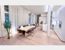 Heller, moderner Wohnraum mit offener Küche, Esstischbereich und großen Fenstern, die mit weißen, transparenten Vorhängen bedeckt sind. Der Boden besteht aus hellem Holz, und im Raum befinden sich stilvolle Möbel sowie eine Topfpflanze in der Ecke.