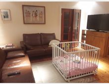 Wohnzimmer mit braunen Ledersofas, einem weißen Laufstall mit rosa Matratze, einem Holzschrank mit Fernseher darauf und einem Teppich auf Holzboden. An der Wand hängen zwei gerahmte Bilder, und im Hintergrund befindet sich eine Doppeltür mit Glasfenstern.