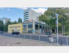 Gelbes zweistöckiges Gebäude mit großen Fenstern, auf dessen Dach der Schriftzug „Albert’s“ steht. Im Hintergrund ist ein mehrstöckiges Wohngebäude zu sehen, umgeben von Bäumen; im Vordergrund befindet sich eine Straße mit parkenden Autos und einem Fahrrad an einem Metallpfosten.