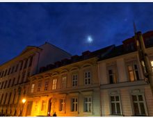 Eine nächtliche Straßenszene mit historischen Stadthäusern, beleuchtet von warmem Laternenlicht. Im Hintergrund ist ein dunkler, wolkenbedeckter Himmel mit einem Lichtschein des Mondes sichtbar.