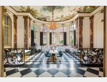 Eleganter Festsaal mit hohen Sprossenfenstern, einer floral bemalten Decke und einem zentralen Kronleuchter. Der Boden besteht aus schwarz-weißen Schachbrettfliesen, und die Wände sind mit Marmorverzierungen und goldenen Details gestaltet. Im Raum stehen gedeckte Tische mit Stühlen sowie ein Buffet entlang der Fensterfront.