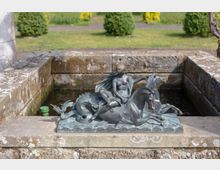 Bronzeskulptur einer Figur, die auf einem mythologischen Pferdewesen mit Fischschwanz sitzt, auf einem steinernen Brunnenbecken platziert, umgeben von einer Gartenlandschaft mit Büschen und gepflegtem Rasen.