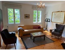 Wohnzimmer mit Holzboden und großen Fenstern, die viel Tageslicht hereinlassen. Zentral stehen ein gelbes Ledersofa und ein Holztisch, ergänzt durch einen gemusterten Sessel und einen schwarzen Stuhl; an der Wand hängen Bilder und ein Kronleuchter hängt von der Decke.