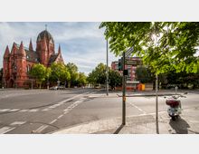 Eine Straßenkreuzung in einer städtischen Umgebung mit einem roten Backsteingebäude im neugotischen Stil, das eine Kirche mit einer Kuppel und einem Kreuz darstellt. Im Vordergrund Straßenschilder, Bäume und ein geparter Motorroller, im Hintergrund Wohnungen und Bäume.