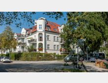 Eckhaus in einer Wohngegend mit heller Fassade und rotem Ziegeldach; Balkone mit Blumenschmuck und umliegende Bäume prägen die Umgebung. Im Vordergrund sind Kopfsteinpflaster, parkende Autos, Fahrräder und Straßenschilder an der Kreuzung „Moltkerstraße“ und „Roonstraße“ zu sehen.