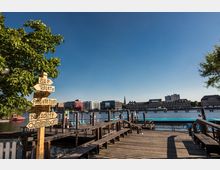 Holzsteg an einem Fluss mit mehreren Sitzbänken und Hinweisschildern, die zu Einrichtungen wie Toiletten, Umkleidekabinen und gastronomischen Angeboten führen. Im Hintergrund moderne und historische Gebäude sowie eine klare, blaue Himmelkulisse.