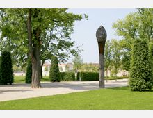 Ein Park mit gepflegten Rasenflächen, großen Bäumen und Hecken. Im Vordergrund steht eine hölzerne Skulptur, während im Hintergrund Gebäude mit hellen Fassaden sichtbar sind.