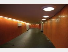 Unterführung mit orangefarbenen Wänden aus Fliesen und einem dunklen Bodenbelag. Die Decke ist mit runden weißen Leuchten ausgestattet, und an den Wänden hängen vereinzelte Schilder.