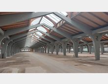 Weitläufige Industriehalle mit einer Dachkonstruktion aus grauen Stahlbalken und Holz. Das Gebäude verfügt über große, lichtdurchlässige Oberlichter und sichtbare Betonstützen, die entlang der Halle in regelmäßigen Abständen angeordnet sind.