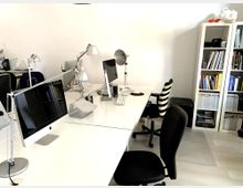 Ein heller Büroraum mit mehreren weißen Schreibtischen, auf denen Computerbildschirme, Schreibtischlampen und Tastaturen stehen. Im Hintergrund ist ein weißes Regal mit Büchern und Ordnern zu sehen.