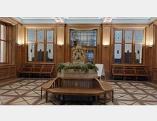 Historischer Innenraum mit holzvertäfelten Wänden und großen Fenstern mit verziertem Glas. Im Zentrum steht ein steinerner Brunnen mit einer Skulptur, die von grünen Pflanzen umgeben ist, umgeben von Holzbänken auf einem dekorativen Bodenmuster.