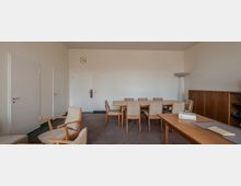 Ein schlichter Konferenzraum mit einer Holzsitzgruppe aus einem langen Tisch und mehreren gepolsterten Stühlen. Im Hintergrund befinden sich eine Uhr an der Wand, zwei Türen und ein Holzschrank, rechts vorne ein weiterer Tisch mit Akten und einer Tischleuchte.