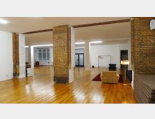 Weitläufiger Innenraum mit Holzboden, Sichtmauerwerk aus Backstein und weißen Wänden. Der Raum verfügt über wenige Möbel, darunter Sitzgelegenheiten, einen roten Teppich, sowie eine Stehlampe; im Hintergrund ist ein Glastüreingang sichtbar.