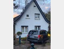 Ein weißes Einfamilienhaus mit steilem, schwarzem Satteldach, zwei kleinen Fenstern im Obergeschoss und einem großen Fenster im Erdgeschoss. Vor dem Haus steht ein dunkler Van auf einer gepflasterten Einfahrt, umgeben von kleinen Büschen und einem beleuchteten Gartenpfosten.