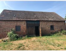 Backsteingebäude mit Satteldach aus dunklen Ziegeln, teilweise beschädigt, sowie zwei kleinen Fenstern und einem hölzernen Tor, das leicht geöffnet ist. Der Boden davor ist grasbewachsen, mit einigen Buschwerkresten am Gebäude.