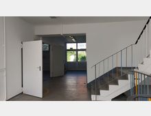 Treppenhaus mit metallenen Geländern, einer offenen weißen Tür auf der linken Seite und einem Durchgang, der zu großen Fenstern mit Blick ins Freie führt. Der Boden besteht aus Beton, und die Wände sind weiß gestrichen.