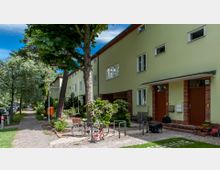 Reihenhäuser mit hellgrünen Fassaden, braunen Türen und roten Klinkerrahmen, umgeben von Bäumen und kleinen Vorgärten; im Vordergrund stehen Fahrräder und Kinderfahrzeuge auf einem gepflasterten Weg. Eine gepflasterte Straße mit parkenden Autos verläuft entlang der Häuser.